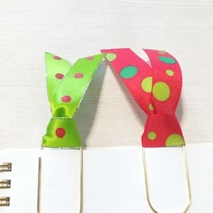 May include: Two gold metal bookmark clips with red and green polka dot ribbon bows. The ribbons are tied in a knot at the top of the clips.