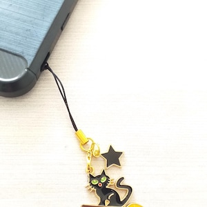 May include: A black cat charm with green eyes and a yellow broom, attached to a black phone strap. A black star charm hangs above the cat. The charm is connected to a black phone case.
