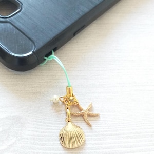 May include: A gold charm with a seashell, starfish, and pearl hanging from a light green string. The charm is attached to a black smartphone case.