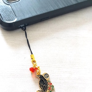 May include: A black crow charm with red and green flowers, a gold charm with a symbol, and a red bead on a black string phone charm.