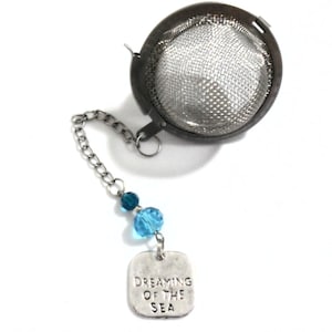 May include: Silver tea infuser with a chain and blue glass beads. The charm on the chain reads "Dreaming of the Sea".