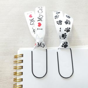 May include: Two black metal bookmark clips with white ribbon attachments. One ribbon has black paw prints and the other has the text "I love My Cat" with a red heart.