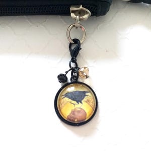 Puede incluir: Una bolsa de cuero negro con cremallera y un dije de metal negro adjunto. El dije presenta un cabuj&oacute;n redondo de vidrio con un cuervo negro y un fondo amarillo.