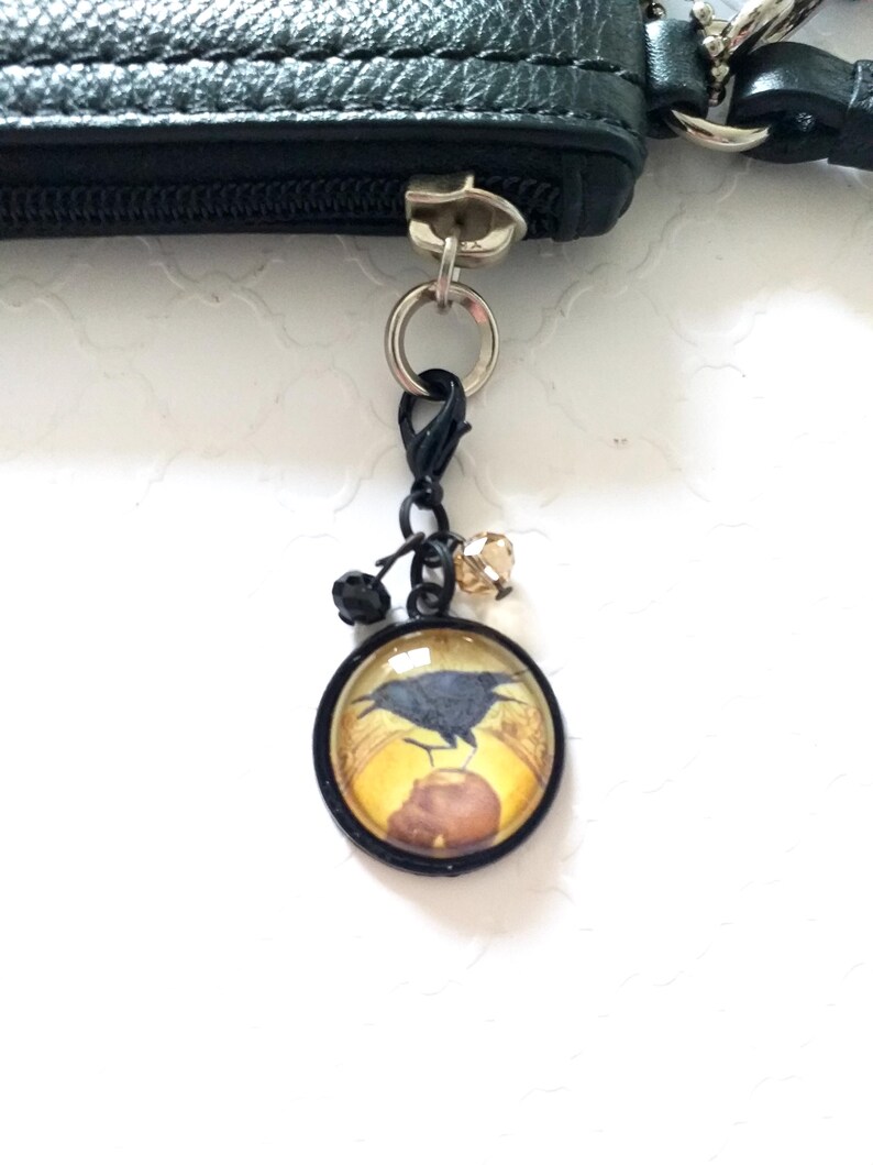 Puede incluir: Una bolsa de cuero negro con cremallera y un dije de metal negro adjunto. El dije presenta un cabuj&oacute;n redondo de vidrio con un cuervo negro y un fondo amarillo.