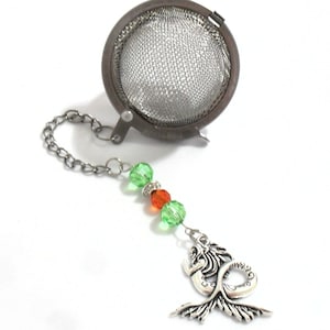 Peut inclure: Infuseur à thé en acier inoxydable avec une chaîne et un charm en forme de sirène. Le charm est orné de perles vertes et oranges. L'infuseur est conçu pour le thé en vrac.