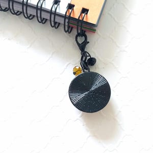 Puede incluir: Marcap&aacute;ginas de metal negro con un disco redondo negro texturizado y una peque&ntilde;a cuenta transparente de color &aacute;mbar. El marcap&aacute;ginas est&aacute; unido a un cuaderno de espiral.