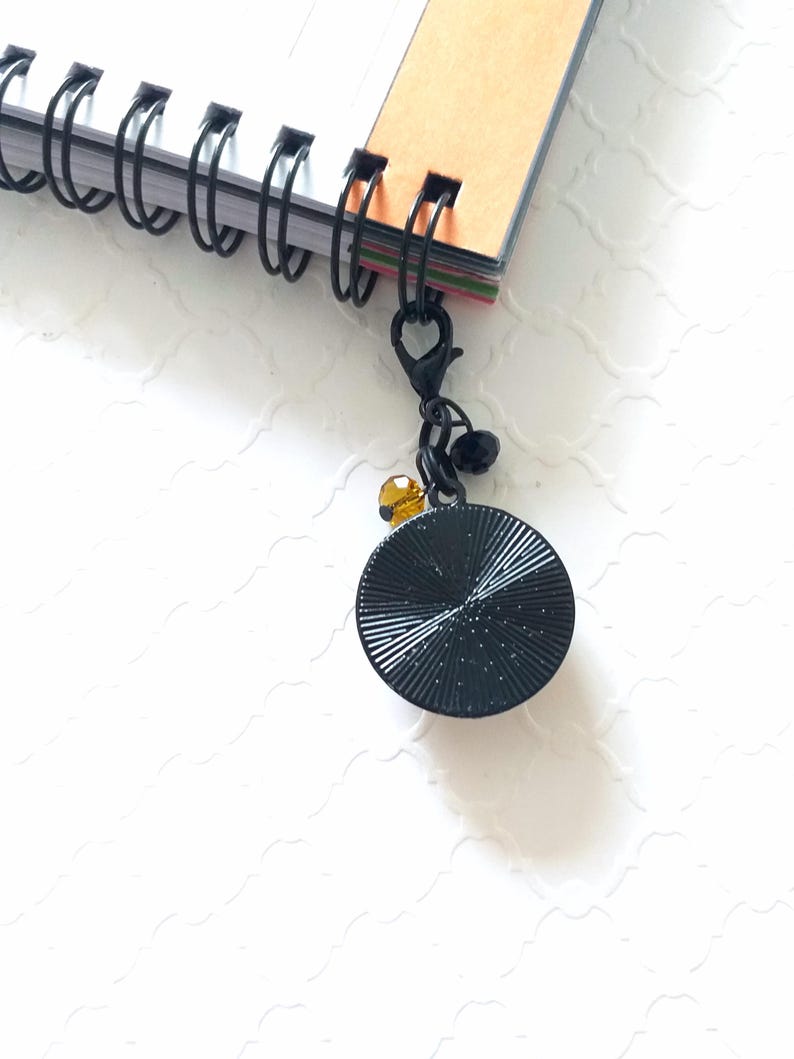 Puede incluir: Marcap&aacute;ginas de metal negro con un disco redondo negro texturizado y una peque&ntilde;a cuenta transparente de color &aacute;mbar. El marcap&aacute;ginas est&aacute; unido a un cuaderno de espiral.