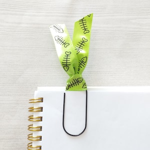 May include: A green ribbon bookmark with a black fishbone pattern, attached to a black metal bookmark clip. The bookmark is on top of a white notebook with gold spiral binding.