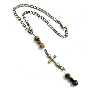 May include: A necklace featuring a bronze-toned metal chain with a lobster clasp, accented by three brown tiger eye beads above a bronze-toned metal cross pendant, followed by two additional brown tiger eye beads at the bottom of the cross.