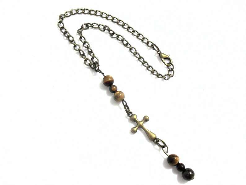 May include: A necklace featuring a bronze-toned metal chain with a lobster clasp, accented by three brown tiger eye beads above a bronze-toned metal cross pendant, followed by two additional brown tiger eye beads at the bottom of the cross.