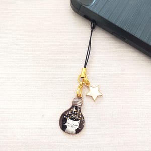 May include: A black lightbulb charm with a white cat inside, surrounded by gold glitter. The charm is attached to a gold chain with a white star charm.