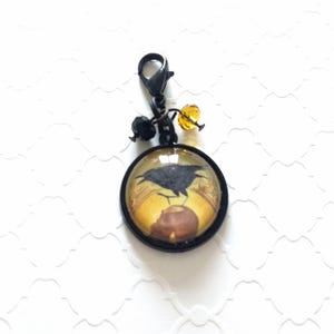 Puede incluir: Un charm de metal negro con un cabuj&oacute;n redondo de vidrio que presenta un cuervo negro posado en una linterna. El charm tiene un cierre de metal negro y est&aacute; adornado con cuentas negras y &aacute;mbar.