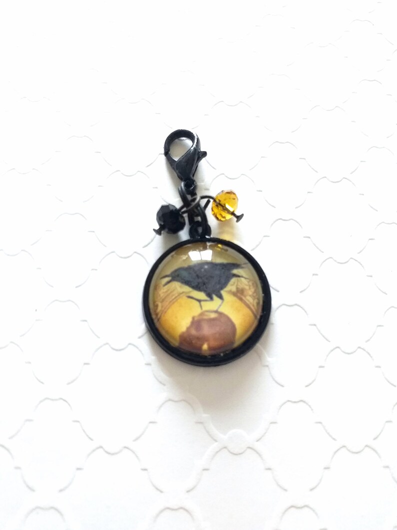 Puede incluir: Un charm de metal negro con un cabuj&oacute;n redondo de vidrio que presenta un cuervo negro posado en una linterna. El charm tiene un cierre de metal negro y est&aacute; adornado con cuentas negras y &aacute;mbar.