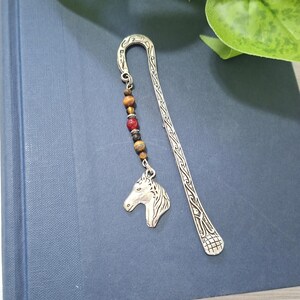 Könnte beinhalten: Ein silberfarbenes Lesezeichen aus Metall mit einem komplizierten eingravierten Muster entlang der Länge. Am oberen Ende befindet sich eine hängende Kette aus vier braunen Tigeraugenperlen, einer roten Perle und einem silbernen Pferdekopfanhänger.
