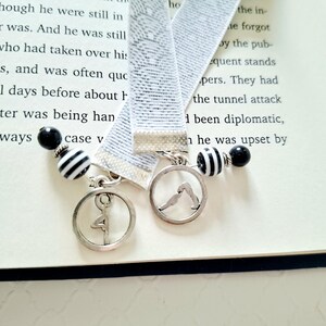 May include: Two elastic bookmarks featuring a light gray woven pattern. Each bookmark is finished with a silver-toned metal clasp, a circular charm depicting a yoga pose, and a decorative bead strand with black and white striped and solid black beads.
