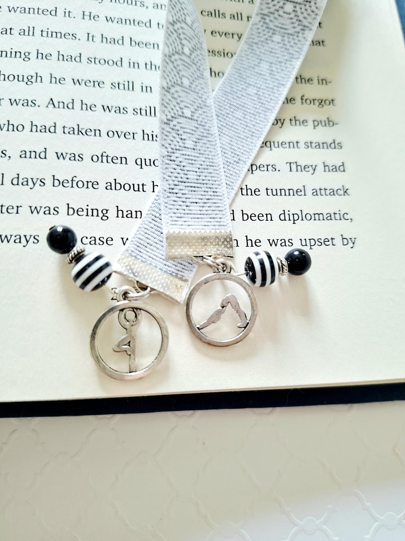 May include: Two elastic bookmarks featuring a light gray woven pattern. Each bookmark is finished with a silver-toned metal clasp, a circular charm depicting a yoga pose, and a decorative bead strand with black and white striped and solid black beads.