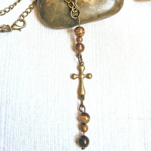 May include: A necklace featuring a bronze-toned metal chain and a central pendant arrangement. The pendant consists of a bronze cross charm flanked by several round, brown, and tan tiger eye beads, suspended in a vertical line from the chain.