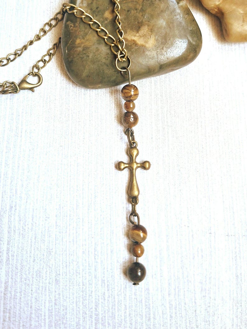 May include: A necklace featuring a bronze-toned metal chain and a central pendant arrangement. The pendant consists of a bronze cross charm flanked by several round, brown, and tan tiger eye beads, suspended in a vertical line from the chain.