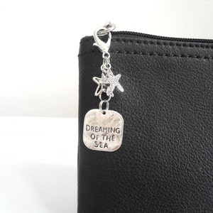May include: A silver charm with a lobster clasp, two starfish charms, and a rectangular tag that reads "DREAMING OF THE SEA." The charm is attached to a black leather pouch.