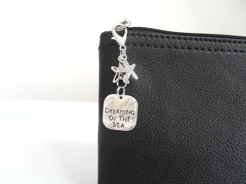 May include: A silver charm with a lobster clasp, two starfish charms, and a rectangular tag that reads "DREAMING OF THE SEA." The charm is attached to a black leather pouch.