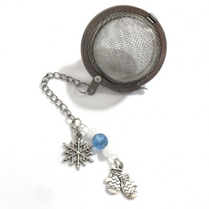Op de afbeelding: Zilveren thee-ei met een ketting en bedels. De bedels zijn een sneeuwvlok en een paar wanten. Het thee-ei is rond en heeft een gaasontwerp.