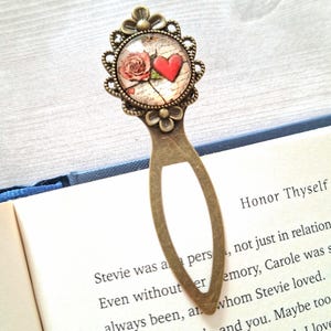May include: A vintage-style metal bookmark with a decorative top featuring a rose and a red heart on a cream-colored background. The bookmark is inserted into a book with the text "Honor Thyself".