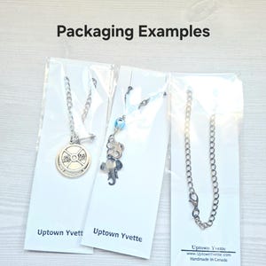 May include: Three silver-toned necklaces displayed on white backing cards labeled Uptown Yvette. The first features a weight plate charm, the second has an elephant and blue bead charm, and the third is a simple chain. Text reads Packaging Examples.