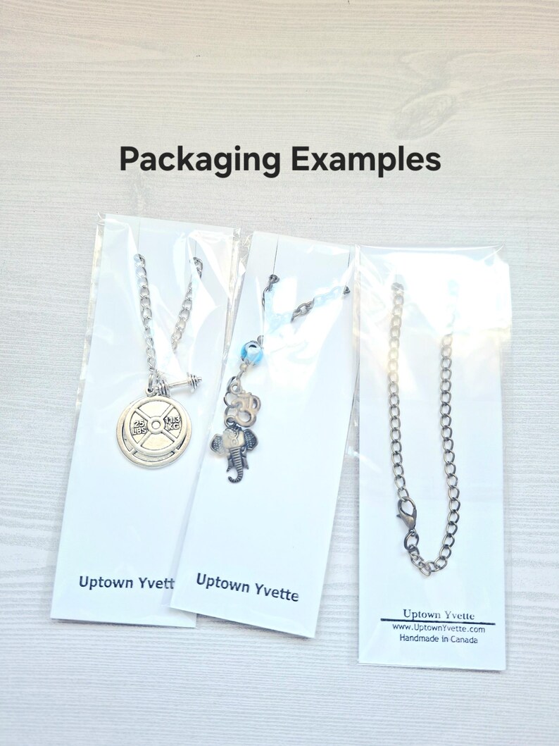 May include: Three silver-toned necklaces displayed on white backing cards labeled Uptown Yvette. The first features a weight plate charm, the second has an elephant and blue bead charm, and the third is a simple chain. Text reads Packaging Examples.
