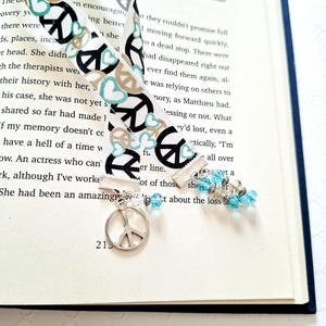 May include: A white fabric bookmark featuring a repeating pattern of black peace signs and light blue hearts. The bookmark is finished with a silver-toned metal peace sign charm at one end and a cluster of light blue and clear beads at the other.