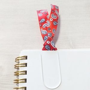 May include: A white spiral bound notebook with a gold coil binding. A red ribbon with a paisley pattern is used as a bookmark.