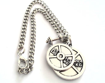 May include: A silver-toned metal necklace featuring a curb chain and a pendant shaped like a miniature barbell with two weight plates. One plate is marked 25 LBS and 11.3 KG, while the other plate is marked 20.4 KG. The necklace includes a lobster claw clasp.