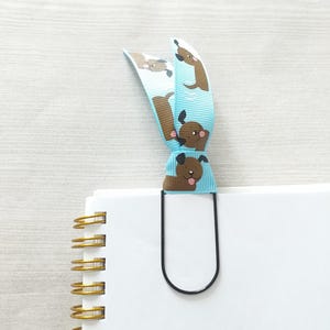 May include: A blue and brown ribbon bookmark with a dog pattern. The bookmark is attached to a black metal bookmark clip.