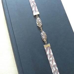 May include: A white and brown fabric bookmark with a silver feather design. The bookmark is attached to a black book.