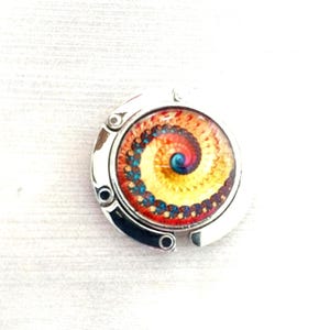 May include: A circular silver-toned metal bag hanger featuring a central glass cabochon with a vibrant spiral pattern. The design displays a gradient of colors transitioning from a deep blue center to yellow, orange, and red, with a dark outer edge.