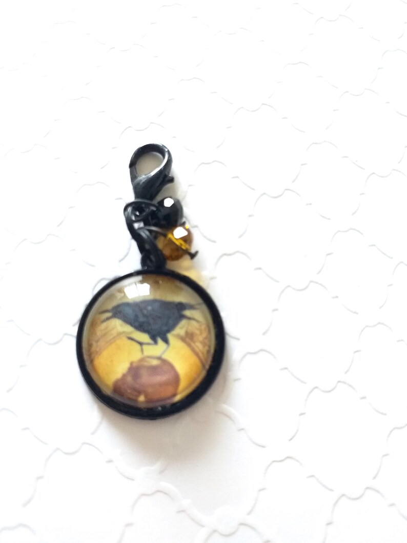 Puede incluir: Un amuleto negro con un cabuj&oacute;n redondo de vidrio que presenta un cuervo negro posado en una rama con un fondo amarillo y marr&oacute;n. El amuleto est&aacute; unido a un cierre de metal negro con cuentas negras y &aacute;mbar.