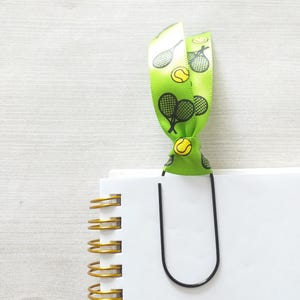 May include: A green ribbon with tennis rackets and tennis balls printed on it is used as a bookmark for a white spiral bound notebook. A black metal paperclip is holding the ribbon in place.