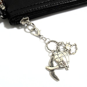 Può includere: Ciondolo argentato con mongolfiera, uccello in volo, sole e nuvola. I ciondoli sono attaccati a un piccolo astuccio nero con cerniera e chiusura argentata. Lo sfondo è bianco.