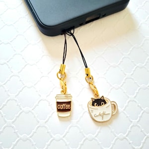 May include: Two phone charms with gold-colored clasps. One charm is a white coffee cup with the word "coffee" in brown. The other is a white mug with a black cat peeking out. The mug has the word "cafe" on it.