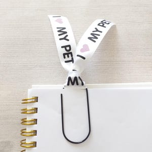 May include: White ribbon bookmark with the text "I love my pet" printed on it. The ribbon is tied around a black metal bookmark clip.