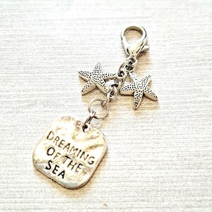 May include: A silver-toned charm with two starfish pendants and a rectangular tag. The tag is engraved with the words "Dreaming of the Sea". The charm is attached to a clasp, suitable for a bracelet or necklace.