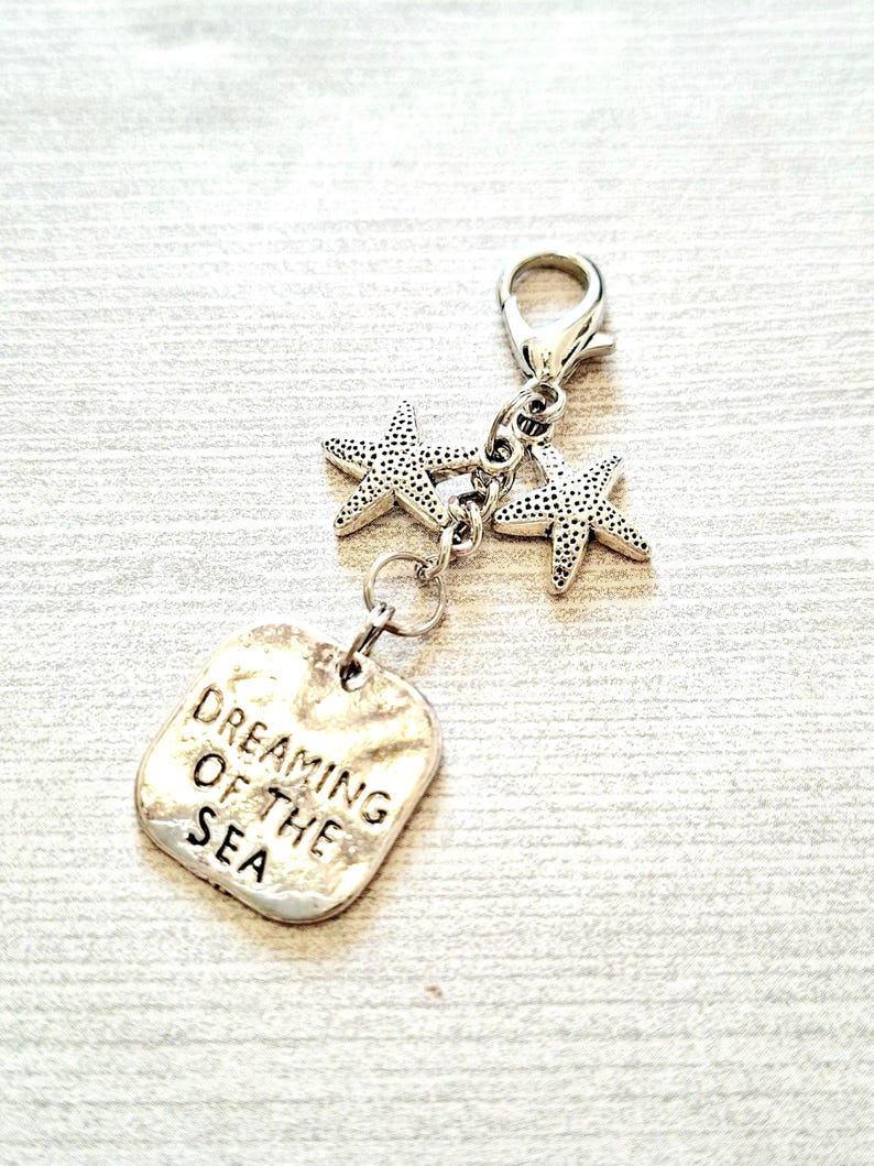 May include: A silver-toned charm with two starfish pendants and a rectangular tag. The tag is engraved with the words "Dreaming of the Sea". The charm is attached to a clasp, suitable for a bracelet or necklace.