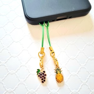 Könnte beinhalten: Zwei Handy-Anhänger an einem schwarzen Smartphone. Ein Anhänger zeigt eine violette Weintraube mit grünem Blatt, der andere eine gelbe Ananas mit grünen Blättern. Beide hängen an dünnen grünen Kordeln mit goldfarbenen Metallbeschlägen.