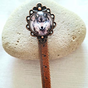 May include: A copper-colored bookmark featuring a detailed illustration of a wolf's face within a floral frame. The bookmark has a ruler design with measurements in inches. The wolf's fur is depicted in shades of gray and brown.
