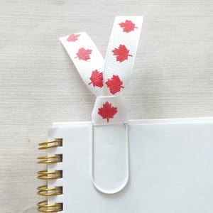 May include: White notebook with a gold spiral binding and a white metal bookmark with a red maple leaf design. The bookmark is tied with a white ribbon with red maple leaf designs.