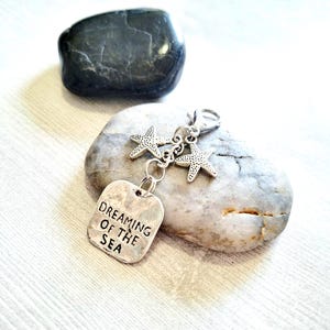 May include: A silver-toned keychain with two starfish charms and a rectangular tag that reads "DREAMING OF THE SEA." The keychain is displayed on a gray stone, with a dark gray stone in the background.