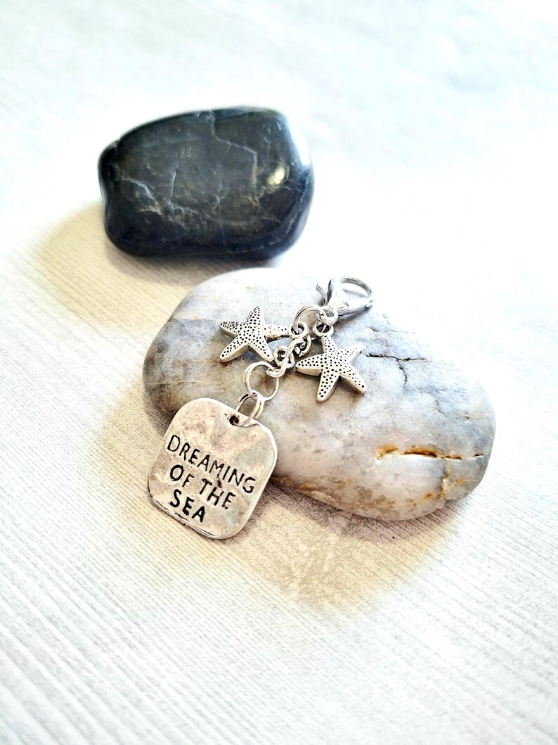 May include: A silver-toned keychain with two starfish charms and a rectangular tag that reads "DREAMING OF THE SEA." The keychain is displayed on a gray stone, with a dark gray stone in the background.