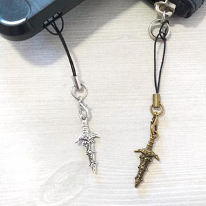 May include: Two silver and bronze sword charms on black cords. The charms are detailed with intricate designs and are attached to phone cases or bags.