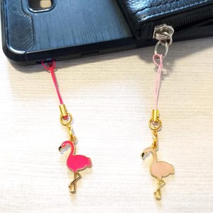 May include: Two flamingo phone charms. One charm is hot pink, the other is light pink. Both charms have gold-colored metal accents and are attached to a pink lanyard. The charms are attached to a black phone and a black wallet.