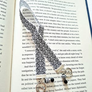 May include: A fabric bookmark featuring a black and white repeating wave pattern. The bookmark is finished with a silver-toned metal clasp, a circular charm depicting a bird in flight, and a dangling bead accent with black and white stripes.