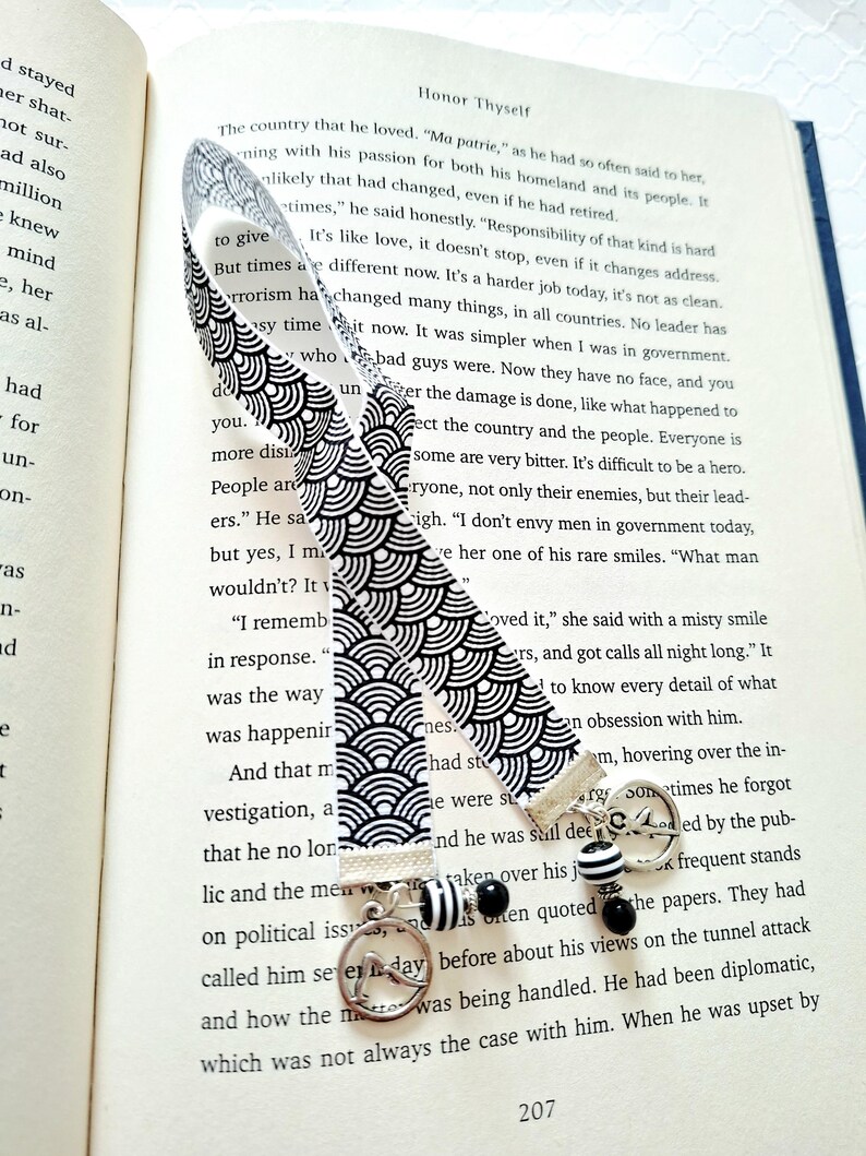 May include: A fabric bookmark featuring a black and white repeating wave pattern. The bookmark is finished with a silver-toned metal clasp, a circular charm depicting a bird in flight, and a dangling bead accent with black and white stripes.